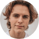 Young person with curly hair smiling at the camera, wearing a white shirt, against a light background.