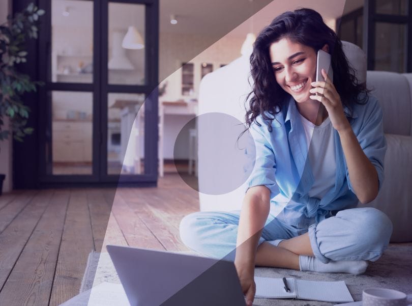 Smiling woman working on a laptop and talking on phone, sitting on floor in bright, modern living room.