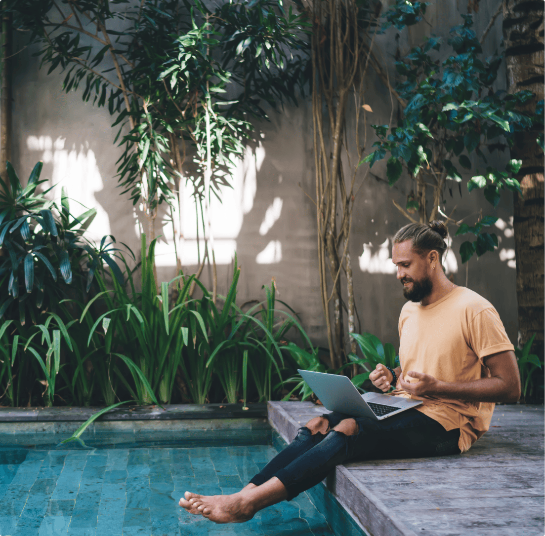 Man sitting by a pool working on a laptop, surrounded by lush greenery, exuding a relaxed digital nomad vibe.