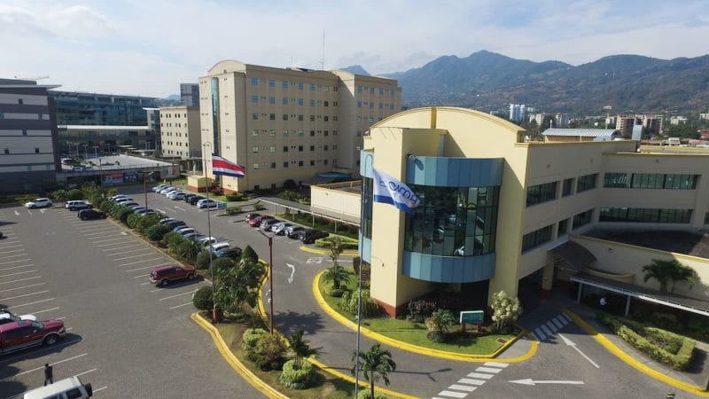 Aerial view of modern hospital complex with parking lot and scenic mountain backdrop.