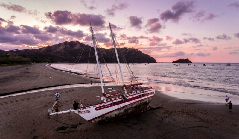 Beached sailboat on a serene coastline at sunset, surrounded by people and lush hills in the background.