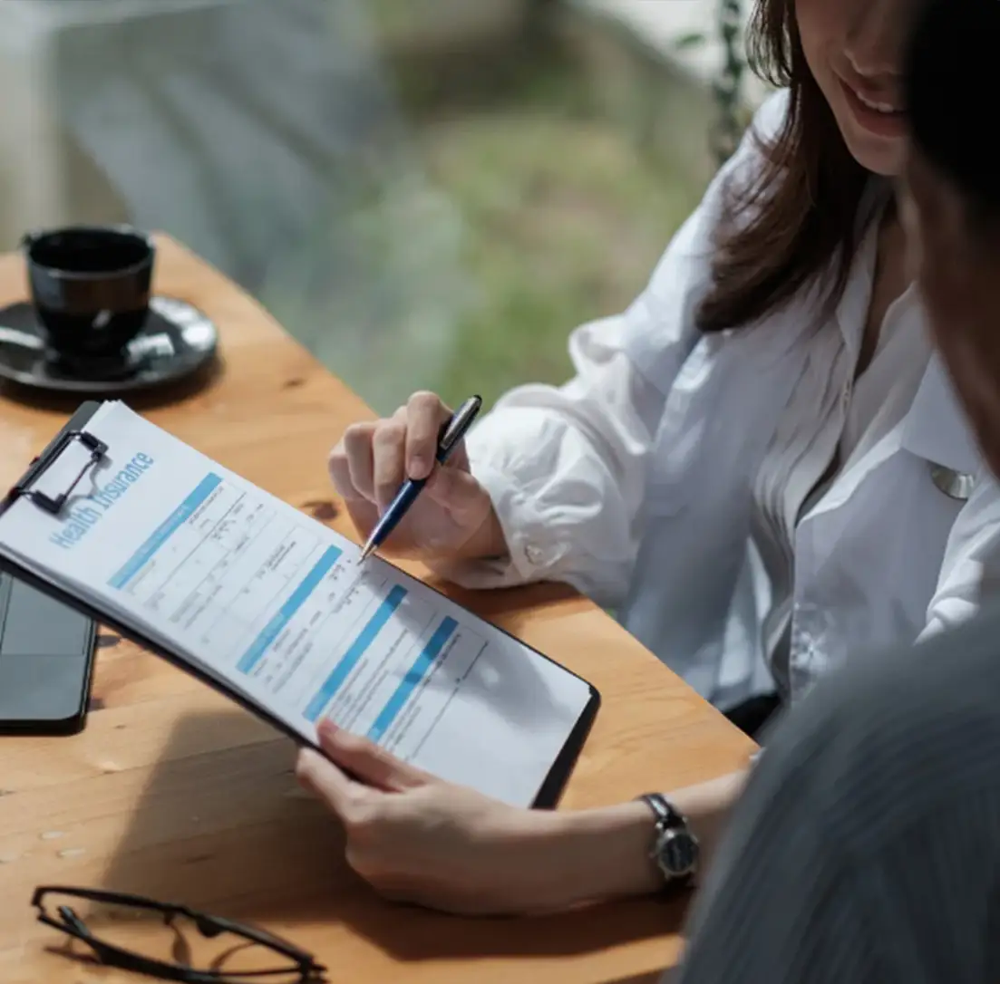 Person discussing health insurance policy details over coffee at a wooden table, using a clipboard.