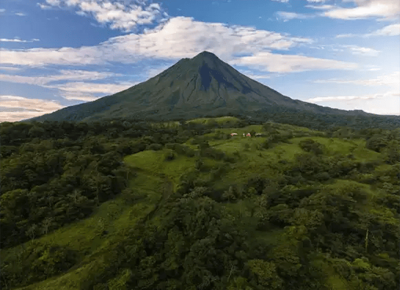 Aerial view of lush green landscape and majestic volcano under a blue sky with clouds.