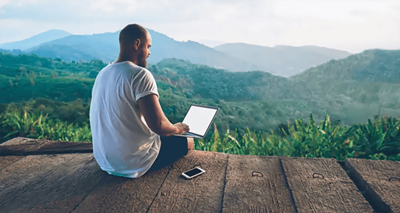 Man working on a laptop with mountain view, representing remote work or digital nomad lifestyle.