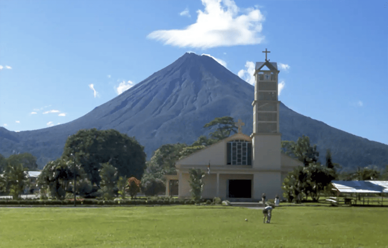 Church and park with majestic Arenal Volcano in Costa Rica's lush landscape under clear blue sky.